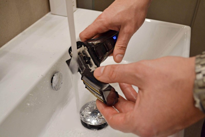 A man is cleaning the head of the electric razor with the water from the tap